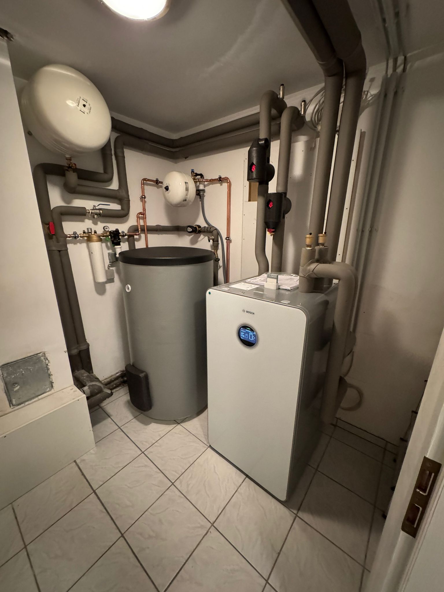 Eine Bosch-Wärmepumpe mit Speicher steht in einem gefliesten Heizungsraum, umgeben von gedämmten Leitungen und Armaturen.